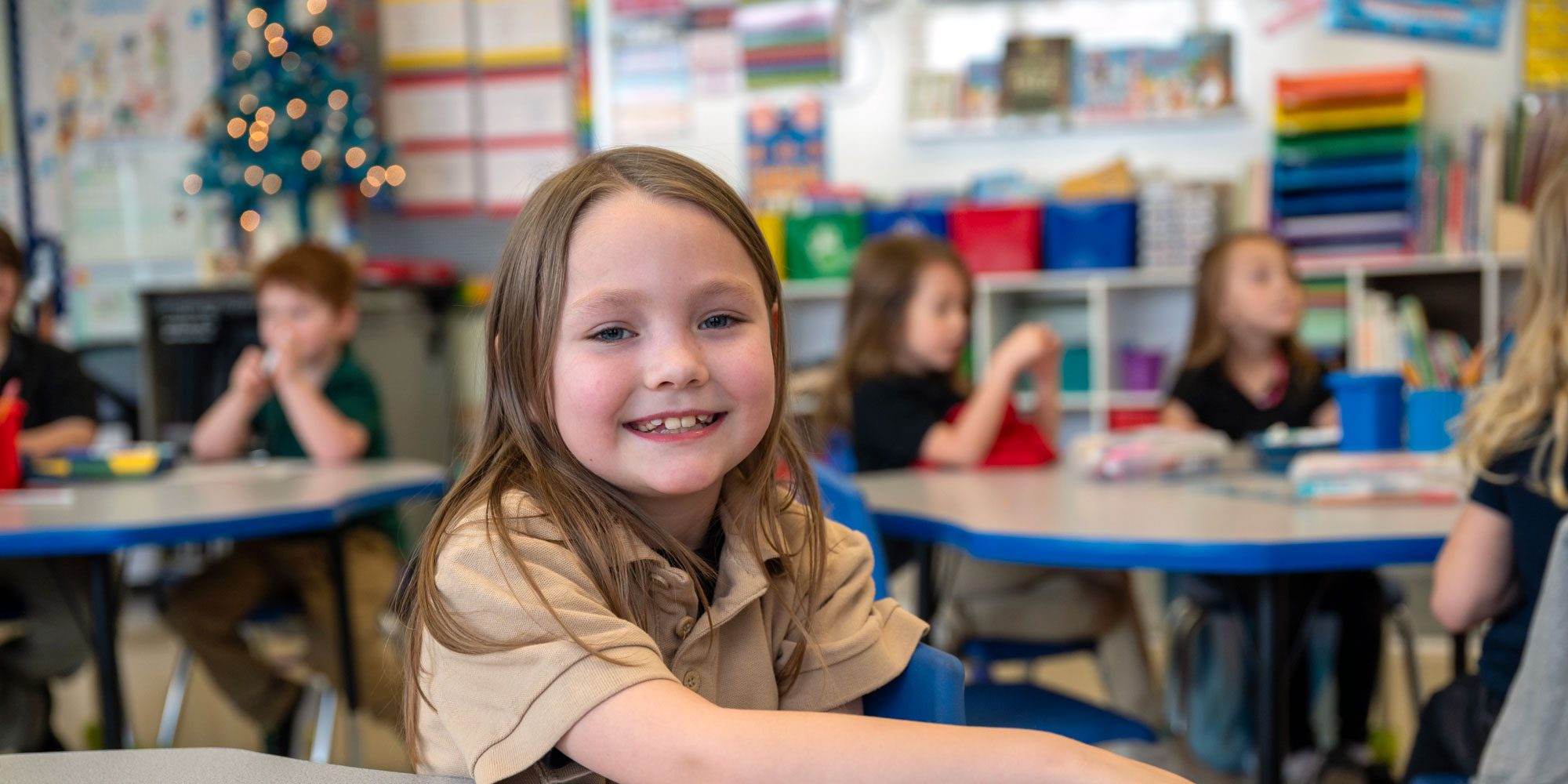 Smiling student at a desk in a classroom with classmates.